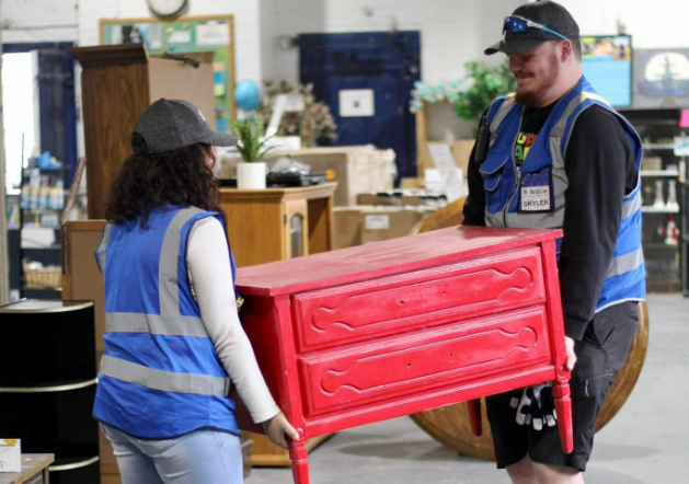 Two ReStore team members in blue safety vests carrying a red painted dresser through the store